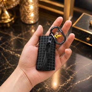 May include: A small black rectangular pouch featuring a white grid pattern and a black zipper closure. Attached to the top is a black carabiner with the text GOBINER and 2M LOCK printed in white, along with a small yellow plastic component.