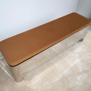 May include: A rectangular bench featuring a smooth, polished silver-tone metal base with rounded corners and a flat, tan-coloured upholstered seat cushion. The piece has a long, low-profile design with a clean, seamless construction.