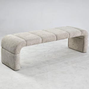 Puede incluir: Un banco tapizado en color beige con una silueta curva y continua, acabado en tela texturizada. El asiento está dividido en cuatro secciones rectangulares acolchadas y la estructura se extiende hacia abajo en dos soportes laterales redondeados.
