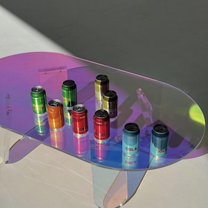 May include: An oval-shaped coffee table with an iridescent, multi-coloured finish holds eight aluminium beverage cans. The cans are arranged in a cluster and feature various colours including green, orange, red, gold, and blue with labels.