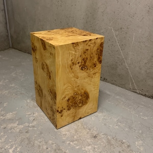 May include: A rectangular pedestal stand crafted from light-coloured wood featuring a prominent, swirling burl grain pattern with dark brown circular clusters throughout the surface. The object has a smooth, polished finish and a solid block structure.