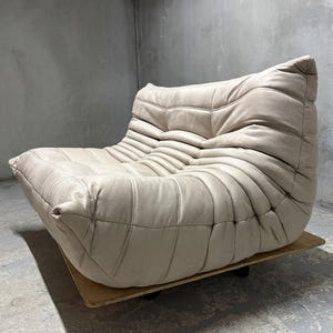 May include: A low-profile, beige leather sofa featuring a distinctive tufted, pleated design with deep horizontal channels across the seat and backrest. The piece is constructed with a soft, rounded silhouette and rests on a flat wooden base.