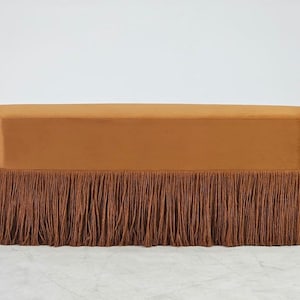 May include: A rectangular bench upholstered in a smooth, solid burnt orange fabric. The base of the bench is adorned with a long, dense fringe trim in a matching deep brown tone that hangs down to the floor, covering the legs completely.
