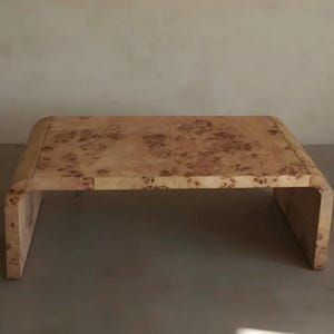 May include: A low-profile coffee table constructed from light-coloured wood featuring a prominent burl grain pattern. The table has a continuous waterfall design with curved edges that transition smoothly from the flat top to the vertical legs.