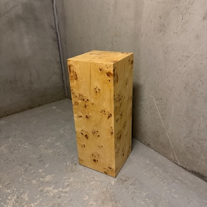 May include: A tall, rectangular pedestal stand crafted from light-coloured wood featuring a prominent burl grain pattern. The structure has a smooth, polished finish with visible natural wood knots and swirling textures across all visible sides.