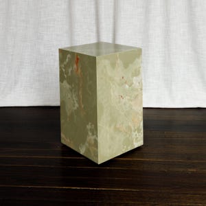 May include: A rectangular pedestal made of light green onyx stone with natural white and orange veining throughout. The stone block features a smooth, polished finish on all visible sides, showcasing the unique mineral patterns of the material.