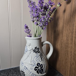 May include: A light gray ceramic pitcher with a handle, featuring embossed blue grape and vine motifs, a figure, and the text "AUS STEIN DEN WEIN" in blue lettering. The pitcher is filled with several stems of purple lavender flowers.