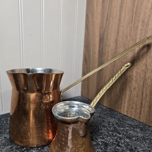 May include: Two copper coffee pots with long brass handles. The larger pot has a smooth finish and a wide pouring spout. The smaller pot features a hammered texture and a decorative, twisted pattern along the length of its brass handle.