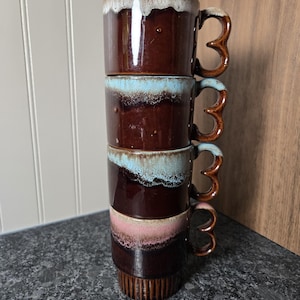 May include: A vertical stack of four ceramic mugs with a dark brown base and a distinctive drip glaze pattern at the rim. Each mug features a unique handle shape and a glossy finish. The top mug has a white drip, the middle two have blue, and the bottom has pink.