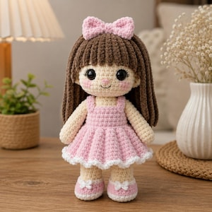May include: A handmade crochet doll featuring brown yarn hair with a pink bow, large black safety eyes, and rosy cheeks. The doll wears a light pink sleeveless dress with a white ruffled hem and matching pink shoes with small white bows.