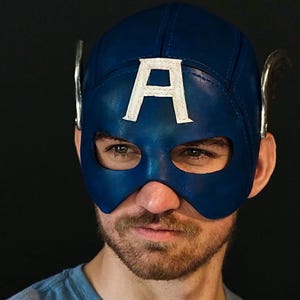 May include: A blue leather mask featuring a prominent white letter A on the forehead and silver-colored wing accents on the sides. The mask covers the upper portion of the face with cutouts for the eyes and is constructed with visible stitching.