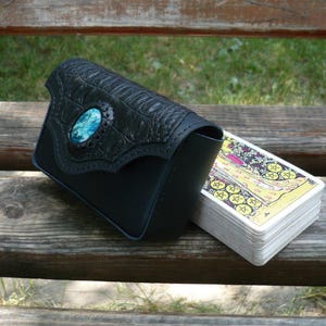 Könnte beinhalten: Eine schwarze Ledertasche mit einer Klappe in Krokodilleder-Optik und einem zentralen ovalen Türkis-Inlay, platziert neben einem Tarotkartendeck mit einer detaillierten Illustration einer Figur zwischen goldenen Münzen und Ranken.