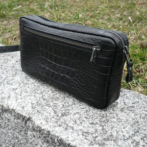 Op de afbeelding: Een rechthoekige zwarte clutch met een getextureerd krokodillenleerpatroon. De tas is voorzien van een horizontaal ritsvak aan de voorzijde, een hoofdritssluiting aan de bovenzijde met een metalen trekker en een bijpassende zwarte polsband.