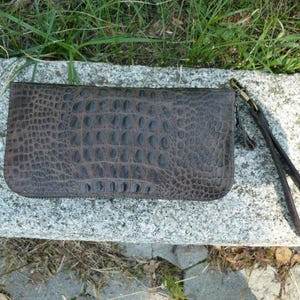 Op de afbeelding: Een rechthoekige donkerbruine portemonnee met een gestructureerd krokodillenleerpatroon. De portemonnee is voorzien van een bijpassende donkerbruine leren polsband die met een metalen sluiting aan de zijkant is bevestigd.