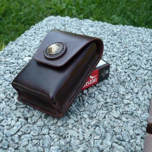 Op de afbeelding: Een donkerbruine lederen etui met een gladde afwerking en een afgeronde klep die wordt vastgezet met een ronde, messingkleurige metalen drukknoop. De etui ligt op een rechthoekige doos met een zwart en rood label met de tekst Winston.