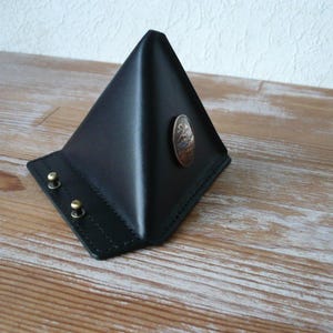 May include: A black leather pyramid-shaped stand featuring a smooth surface, two small brass-coloured metal studs on the front base, and a single circular metallic emblem with an embossed design centred on one of the triangular side panels.