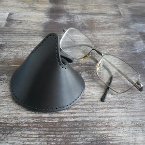 May include: A black leather glasses case with a conical shape and visible stitching along the edges, positioned next to a pair of rectangular spectacles with thin silver metal frames and clear lenses resting on the surface.