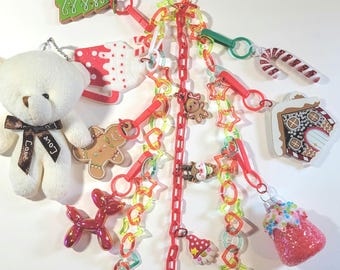 May include: A red chain with various Christmas-themed charms, including a gingerbread house, a candy cane, a snowman, a Christmas tree, a gingerbread man, a red balloon, a white teddy bear with a brown bow, and a red and white ornament. The chain is attached to a gold paperclip.