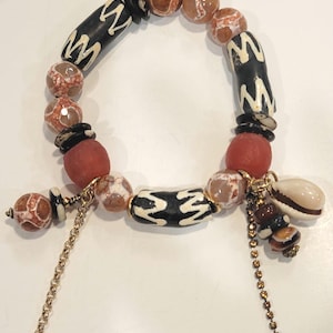 May include: A beaded bracelet with a mix of brown, white, and red beads. The bracelet has a gold chain with a shell charm and a small gold charm.