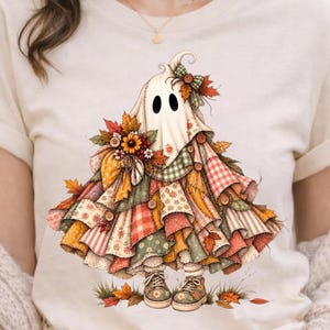 Peut inclure: Un t-shirt couleur crème avec un motif représentant un fantôme portant une robe en patchwork superposé, composée de divers tissus aux motifs automnaux, incluant des carreaux et des fleurs, agrémentée de boutons, de feuilles et d'un nœud vert.