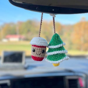 May include: Two crocheted car charms hanging from a car mirror. One charm is a coffee cup with a white lid, brown cup, and red accents. The other is a green Christmas tree with white and yellow accents. Both charms have brown strings.