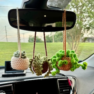May include: Three crocheted hanging plant decorations in a car. The plants are in small, brown pots with cream, brown, and green yarn. The plants are suspended from the rearview mirror by cream and brown yarn.