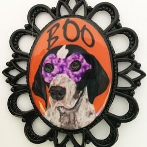 May include: A Halloween-themed oval pendant featuring a dog wearing a purple mask, with the word "BOO" in black letters. The dog is set against an orange background, framed by a black filigree border. A festive accessory.