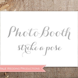 May include: White sign with the text "Photo Booth strike a pose" in a silver cursive font. The sign has a pink banner at the bottom with the text "Fairytale Wedding Productions".