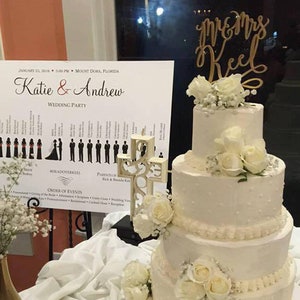 May include: A white wedding cake with white flowers and a gold "Mr. & Mrs. Keel" topper. The cake is on a table with a sign that says "Katie & Andrew Wedding Party" and a list of the wedding party members.