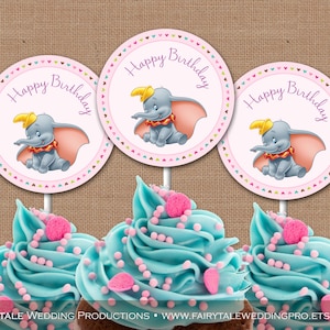 May include: Three blue cupcakes with pink frosting and sprinkles topped with round pink and white cupcake toppers featuring a cartoon image of Dumbo the elephant with the text "Happy Birthday".