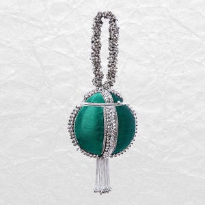 Pode incluir: Uma bolsa esférica de cetim verde esmeralda com adornos de contas metálicas prateadas ao longo das costuras e uma alça de contas a condizer. A bolsa inclui uma borla de franjas metálicas prateadas pendurada no centro da base redonda.