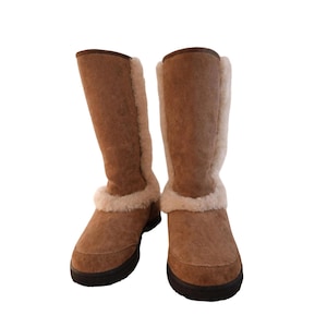 May include: A pair of mid-calf boots in a tan suede finish featuring a thick cream-colored faux fur lining that extends along the front seam and around the ankle. The boots have a rounded toe and a dark brown flat rubber sole for traction.