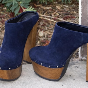 May include: A pair of navy blue suede mules featuring a high wooden platform sole and a thick wooden block heel. The base of the upper is accented with a row of small silver-toned metal studs along the perimeter where the suede meets the wood.