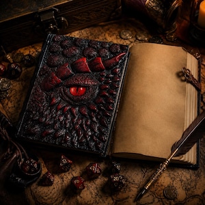 May include: A journal with a textured black and red cover featuring a prominent raised dragon eye design. The book is open to blank, aged-looking pages. A dark feather quill pen rests on the open page, surrounded by several multi-sided dice.