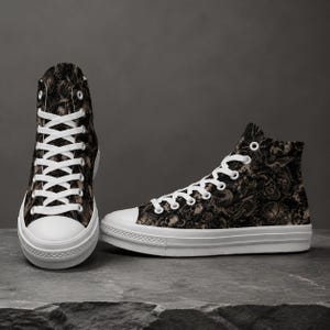 May include: A pair of high-top sneakers featuring a dark, intricate pattern of skulls and roses in shades of black, brown, and cream. The shoes have white rubber toe caps, white rubber soles, and white laces threaded through silver-toned metal eyelets.