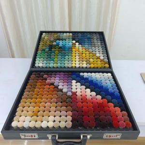 Op de afbeelding: Een zwarte koffer met een grote collectie kleine, ronde, pluizige garen pompons gerangschikt in een raster. De pompons tonen een breed kleurenspectrum, waaronder tinten blauw, groen, geel, bruin, paars, rood en wit.