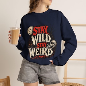 Puede incluir: Una sudadera azul marino de manga larga con un estampado gráfico que incluye el texto "STAY WILD STAY WEIRD" en rojo y blanco roto. El diseño presenta una luna llena, una calavera, la silueta de una casa y ramas de árboles desnudas.