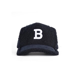Peut inclure: Une casquette de baseball bleu marine foncé fabriquée dans une matière texturée et duveteuse semblable à du sherpa. Le panneau avant présente une grande lettre B majuscule blanche brodée, centrée sur la calotte. La visière est également recouverte du même tissu duveteux bleu marine foncé, créant une apparence uniforme et cohérente sur toute la structure de la casquette.