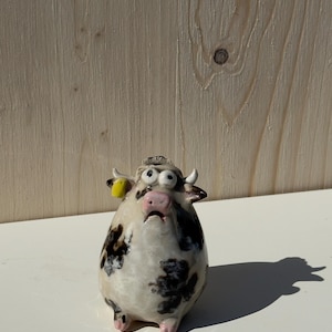 Può includere: Una statuetta di mucca in ceramica dal corpo arrotondato, caratterizzata da una base bianca con macchie irregolari nere e marroni. La mucca ha un'espressione sorpresa con occhi spalancati, un muso rosa, piccole corna e una targhetta gialla.