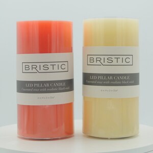 May include: Two LED pillar candles, one in coral and one in cream, with the text "BRISTIC LED PILLAR CANDLE Immersed wax with realistic black wick 6in H x 3 in Dia" on the label.