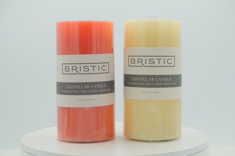 May include: Two LED pillar candles, one in coral and one in cream, with the text "BRISTIC LED PILLAR CANDLE Immersed wax with realistic black wick 6in H x 3 in Dia" on the label.