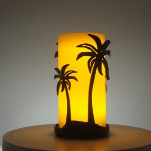 May include: A yellow candle with a black metal palm tree design. The candle is on a wooden surface.