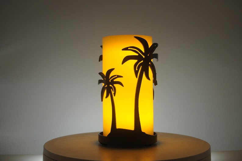 May include: A yellow candle with a black metal palm tree design. The candle is on a wooden surface.