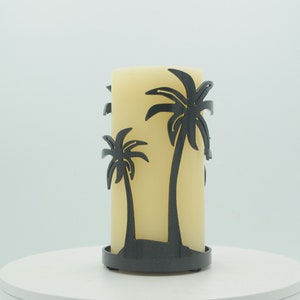 May include: A black metal candle holder with a silhouette of two palm trees. The candle holder is holding a large, off-white pillar candle.