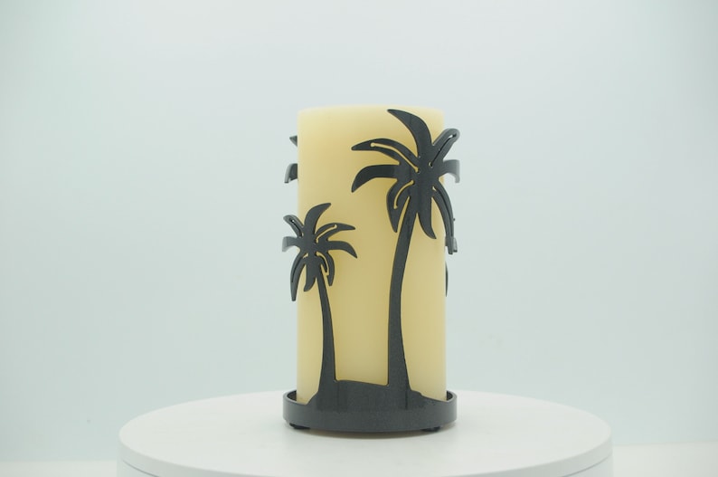 May include: A black metal candle holder with a silhouette of two palm trees. The candle holder is holding a large, off-white pillar candle.