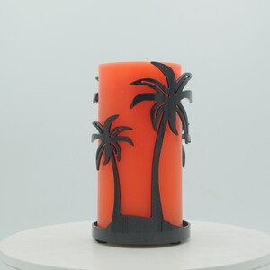 May include: Orange pillar candle with a black metal palm tree design holder. The candle is sitting on a white surface.