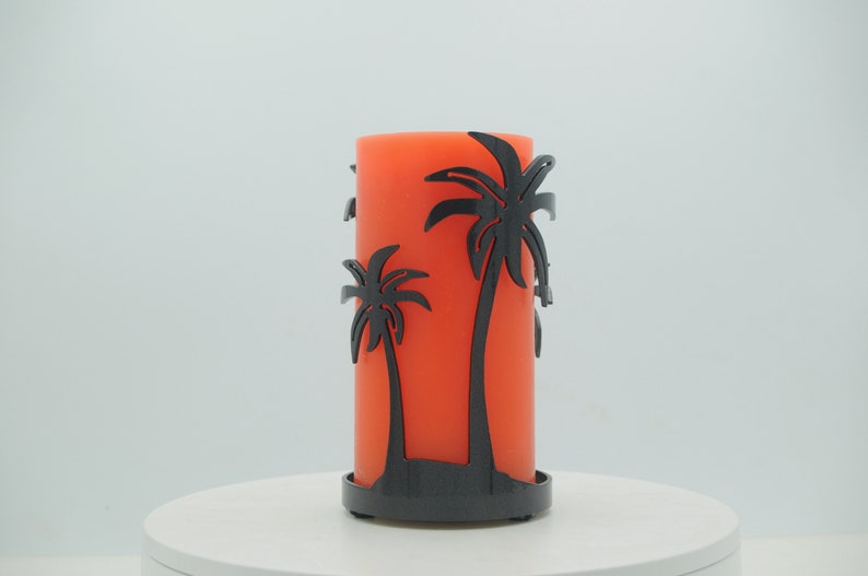 May include: Orange pillar candle with a black metal palm tree design holder. The candle is sitting on a white surface.