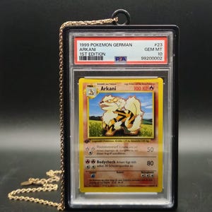 Könnte beinhalten: Eine deutsche 1999er 1st Edition Arkani Pokemon-Karte, bewertet mit PSA Gem Mt 10, eingeschlossen in einer schützenden Kunststoffhülle mit schwarzem Rand. Die Karte hat eine goldfarbene Metallkette an der Oberseite, um sie als Anhänger zu tragen.