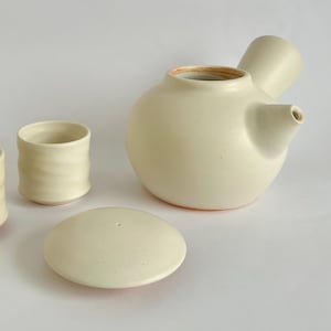 May include: A ceramic tea set consisting of a side-handled teapot with a separate lid and two matching cups. The pieces feature a smooth, off-white matte finish with subtle speckles and a visible terracotta-coloured clay base at the bottom.