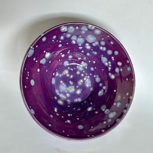 May include: A round ceramic bowl with a deep purple glaze featuring a speckled pattern of light blue and white circular spots. The interior surface is smooth and glossy, displaying a vibrant color gradient that transitions from dark purple to lighter tones.